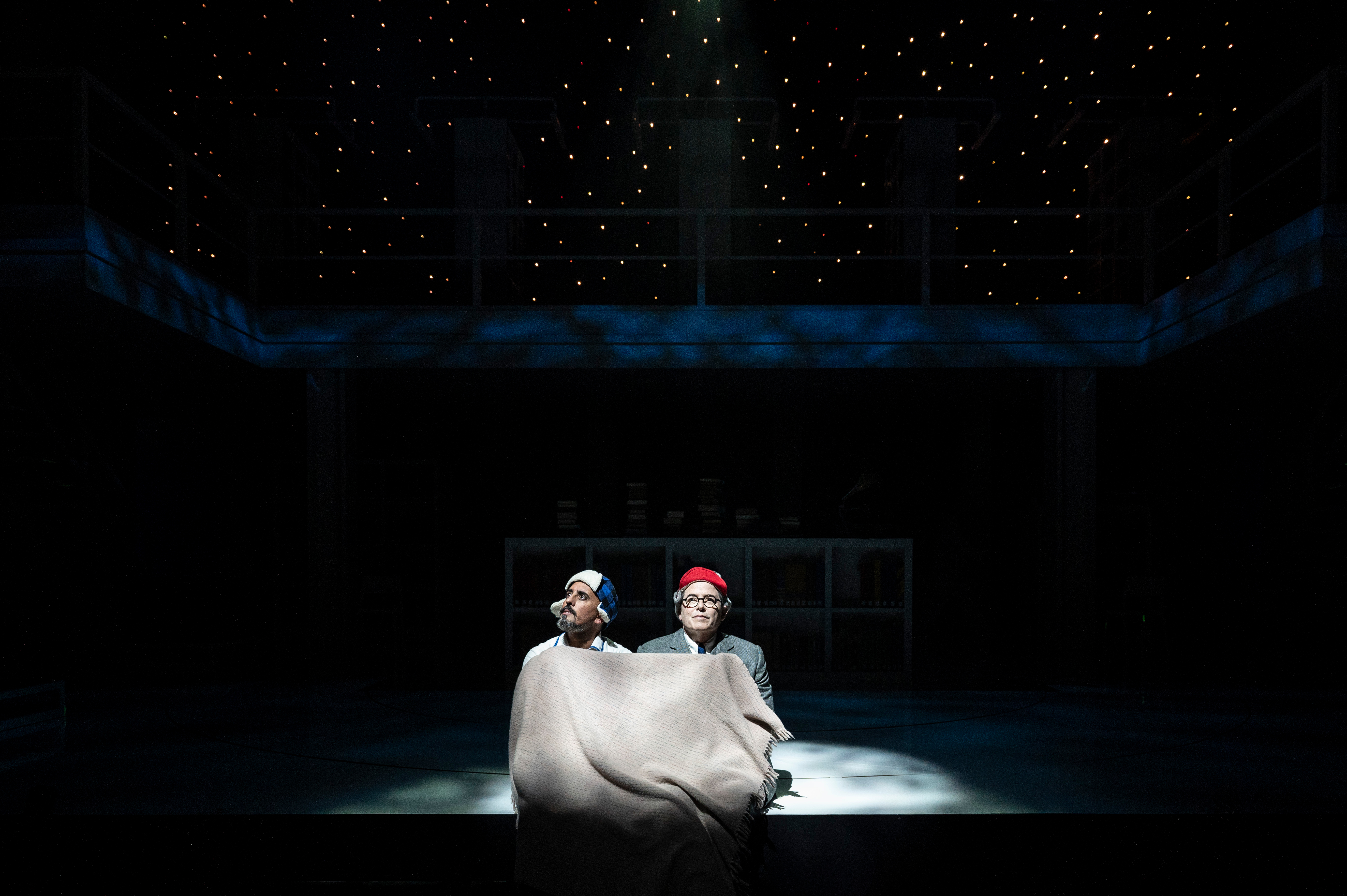 Nehal Joshi and Matthew Broderick beneath a khaki blanket on a dark stage with starlight overhead.