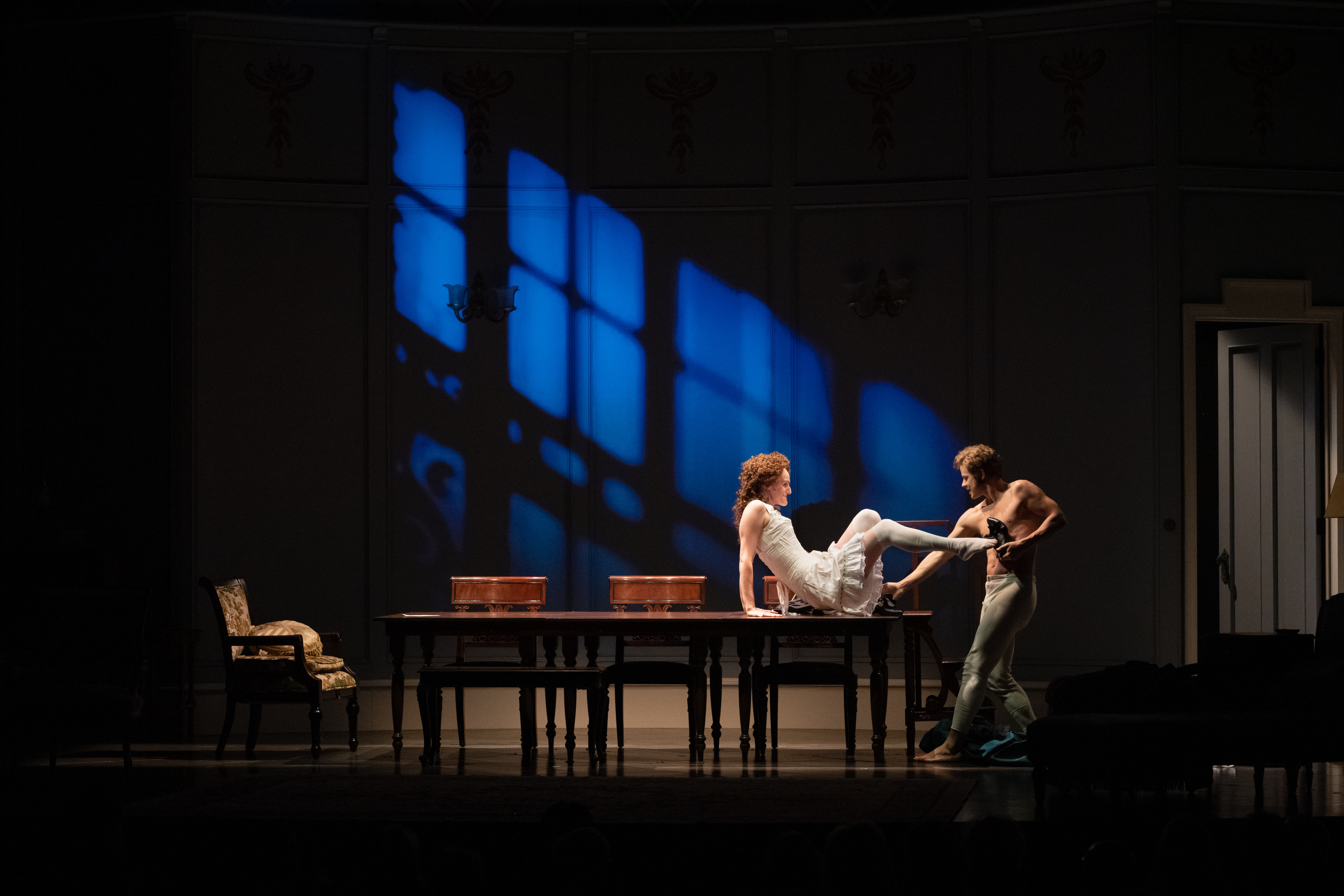 A darkly lit scene featuring a man and woman half dressed, the woman seated atop the table while intensely gazing at the man, standing and equally as focused on her.