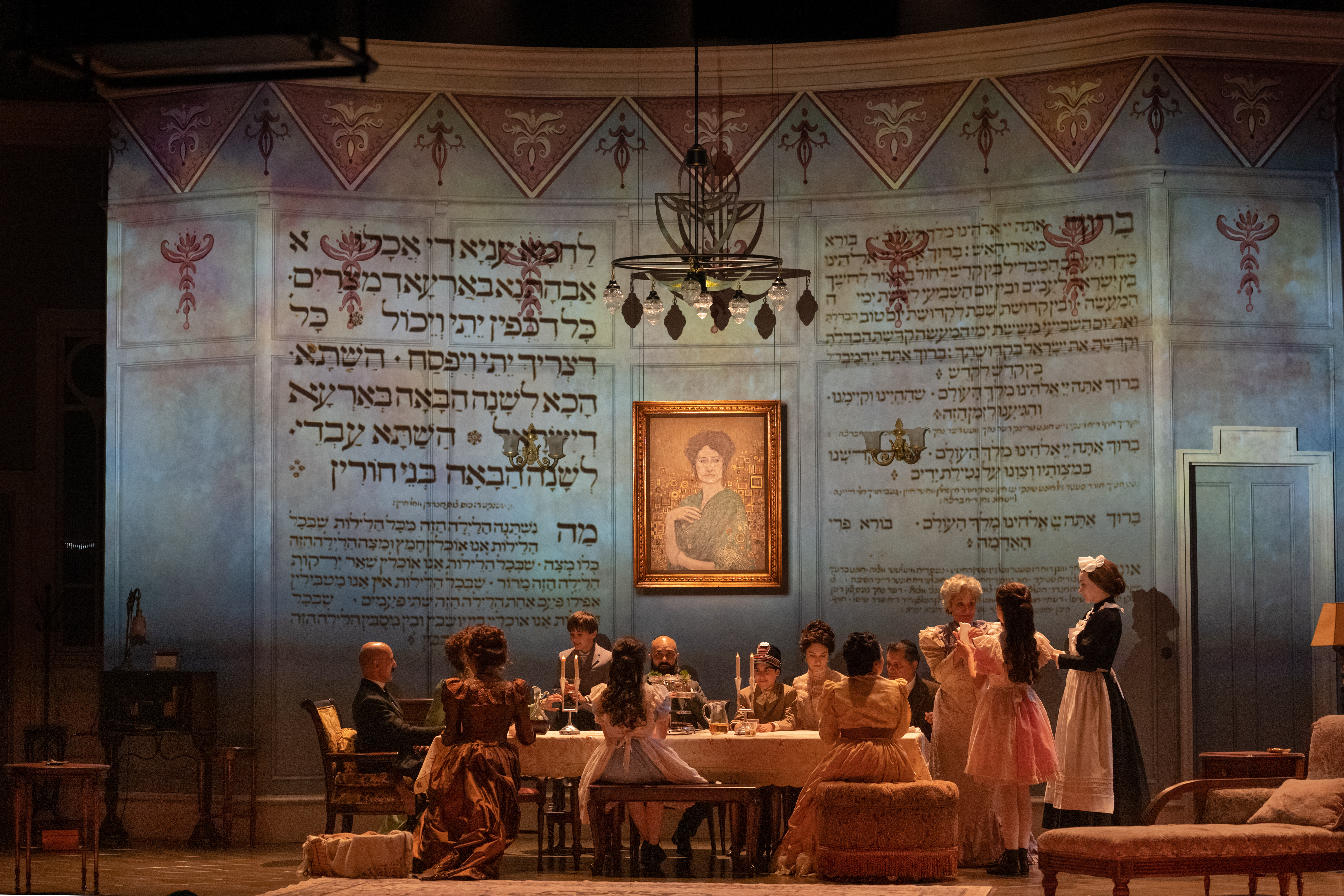 A wide shot of a dinner or seder. Hebrew text is projected onto the back wall of the home.